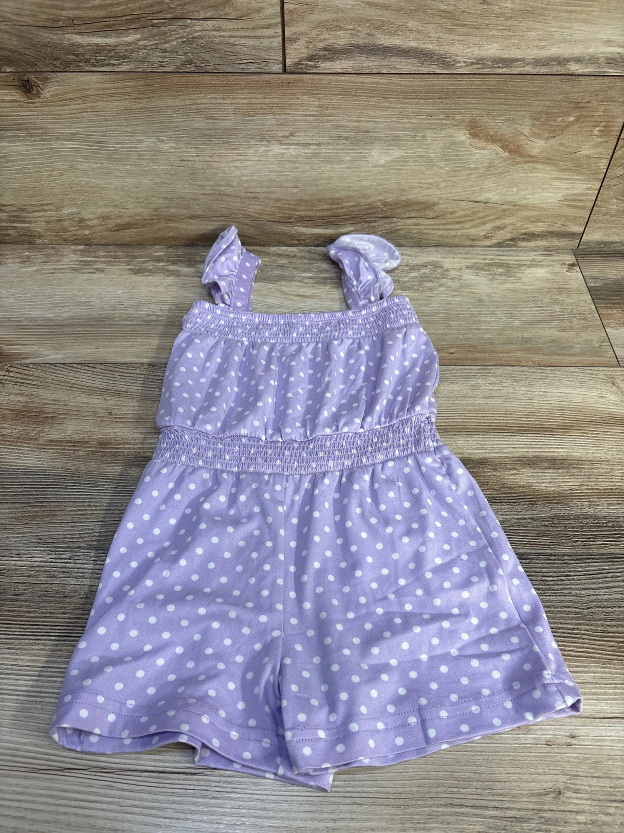 Flat lay of purple polka-dot shortie romper showing ruffled shoulder straps and smocked waist on light wood background