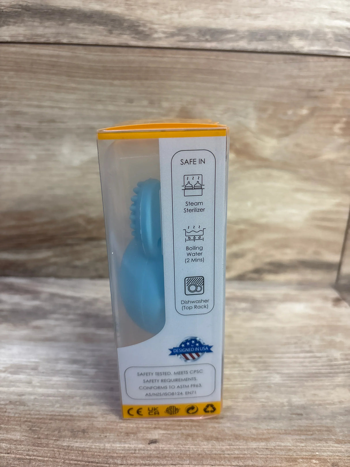 Side view of boxed blue bunny egg teething toy showcasing textured chew ring and sterilization icons on packaging with wooden background