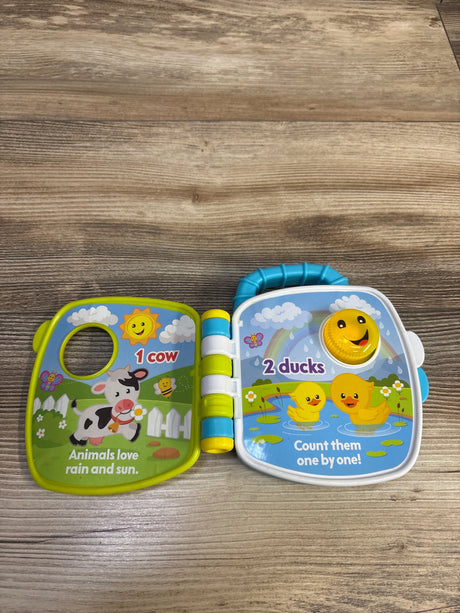 Top-down view of Fisher-Price Counting Animal Friends electronic book open to pages labeled 1 cow and 2 ducks with yellow smiling dial on wood surface