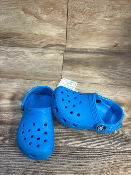 Three-quarter view of blue Crocs Classic clogs showcasing ventilation holes and adjustable heel strap with wood-grain floor background