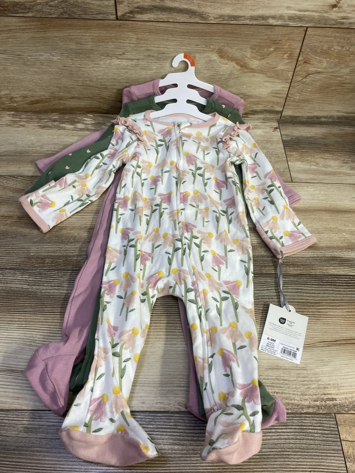 Front view of Cloud Island three pack baby sleepers in pink, green, and floral prints, size 6-9 months, on a wood floor