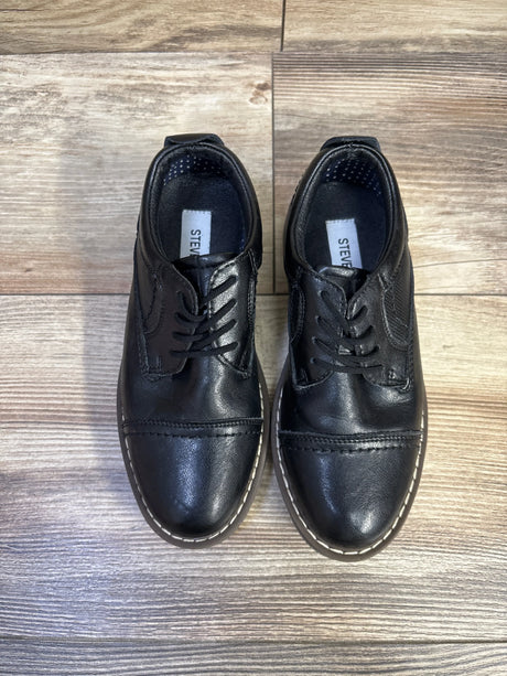 Top-down view of Steve Madden Toliverr toddler black cap-toe oxford shoes showing white stitched welt and lace-up front on wood floor