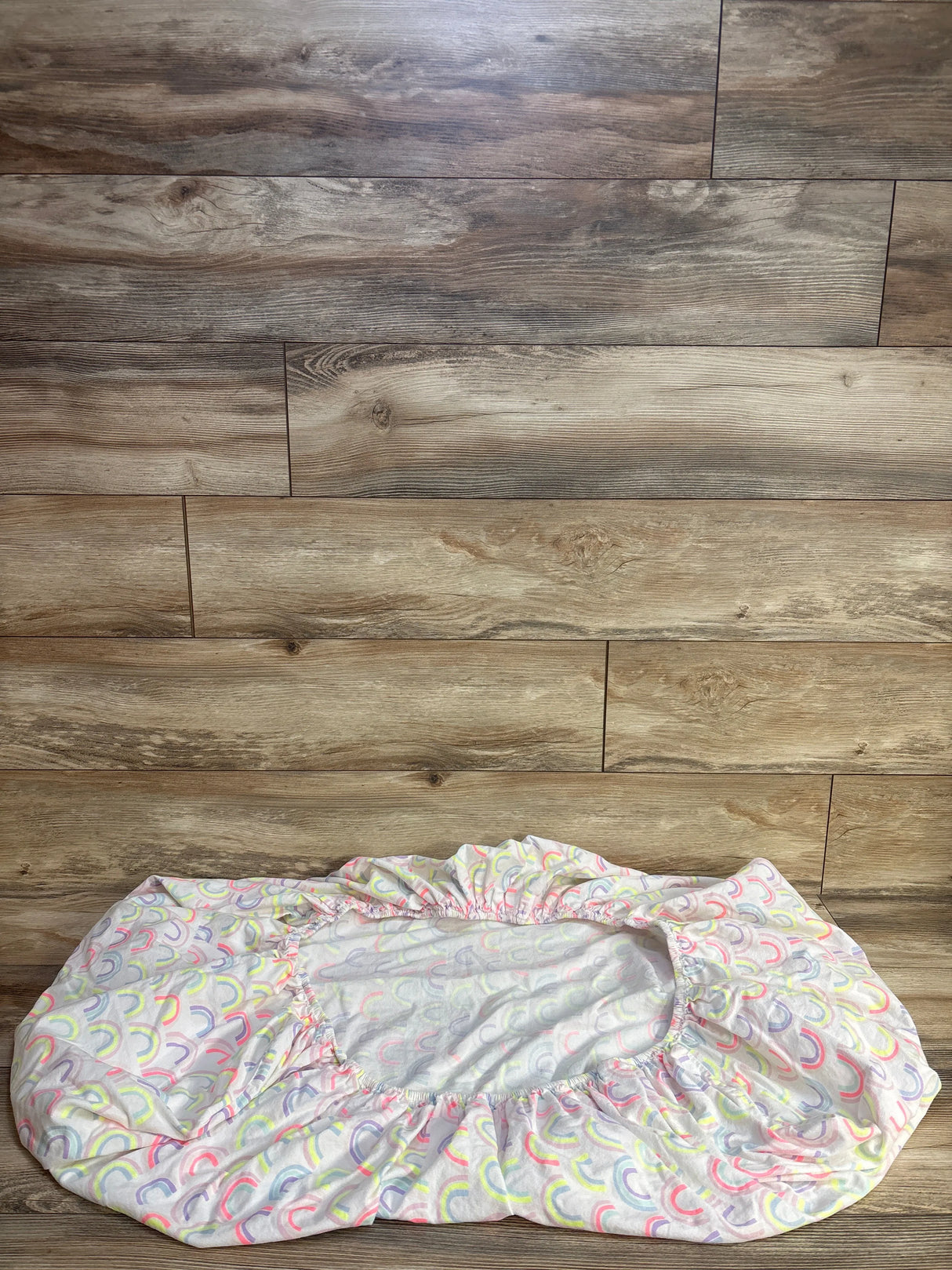 Top-down view of Cloud Island fitted crib sheet showing pastel rainbow arches on white fabric with elastic edges on wood floor