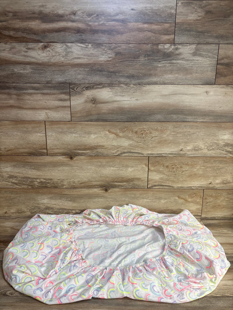 Top-down view of Cloud Island fitted crib sheet showing pastel rainbow arches on white fabric with elastic edges on wood floor