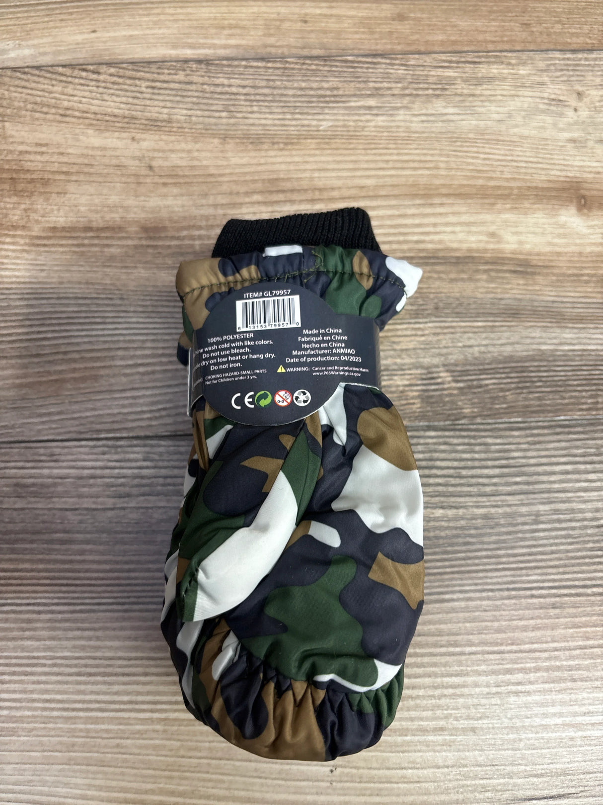 Child's camo pattern winter mitten with black cuff and product information label on wooden surface
