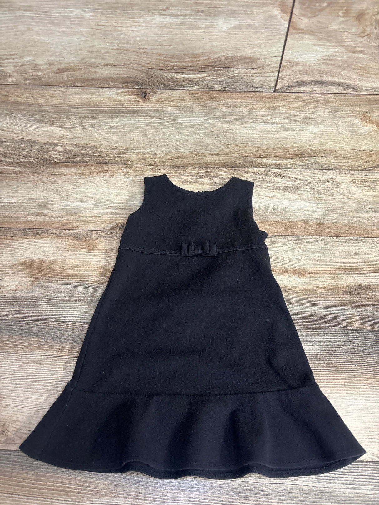 Gently used sleeveless black toddler dress with ruffled hem and small bow on waist, laid on wooden floor
