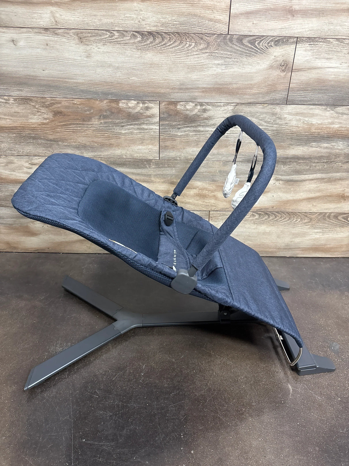 Three-quarter view of Baby Delight Alpine Deluxe Bouncer in quilted indigo showcasing reclined seat with toy bar on wood plank floor
