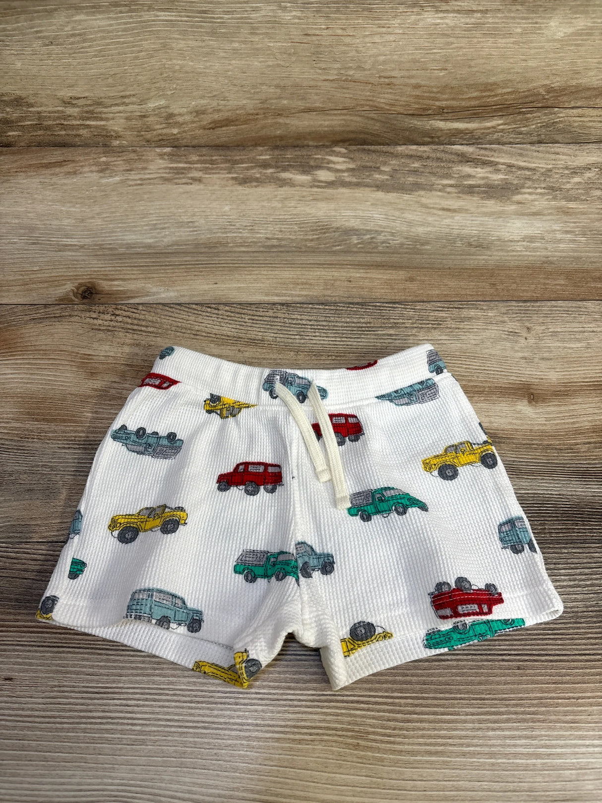 Flat lay of white Old Navy waffle-knit baby shorts with multicolor car print and drawstring waistband on wood background