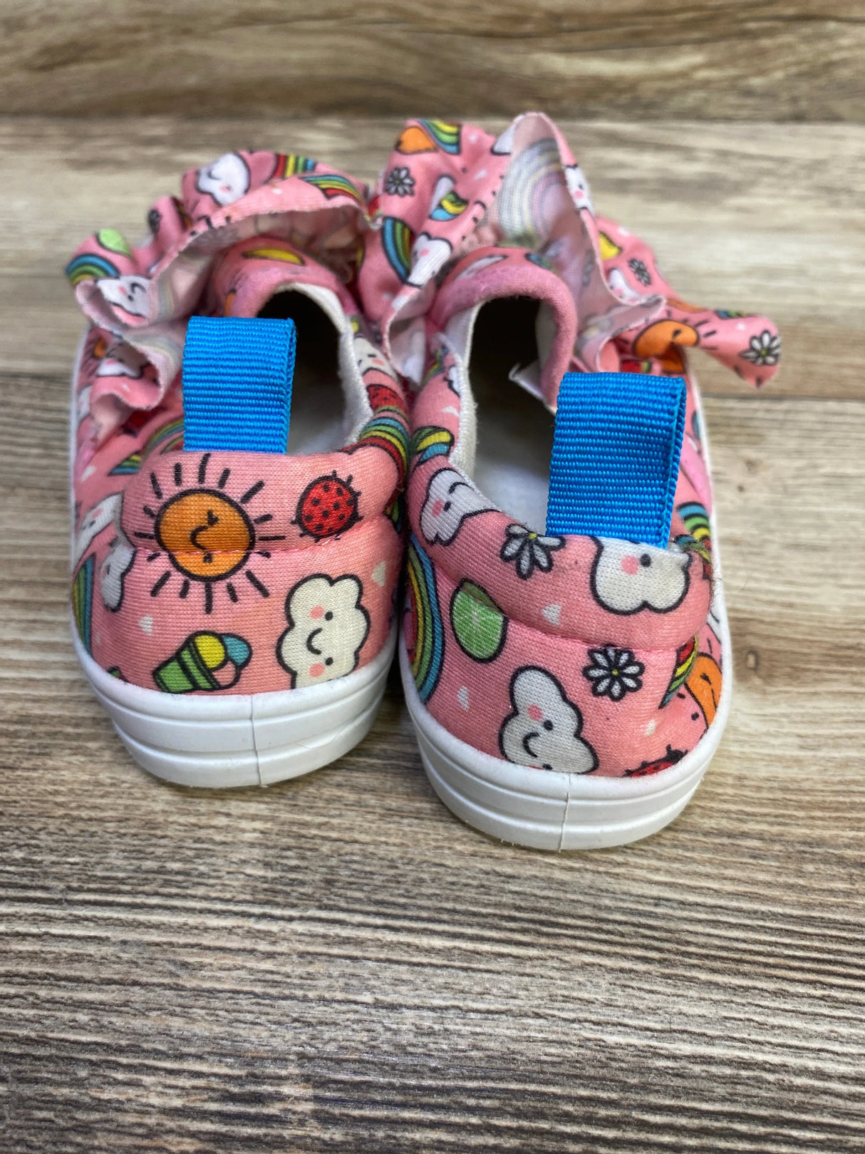 Rear view of Limited Too pink slip-on sneakers with ruffle collar, rainbow and cloud print and blue tabs on wood floor