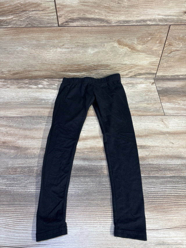 Flat-lay of toddler black leggings (size 2T) showing elastic waistband and slim tapered legs on light wood floor