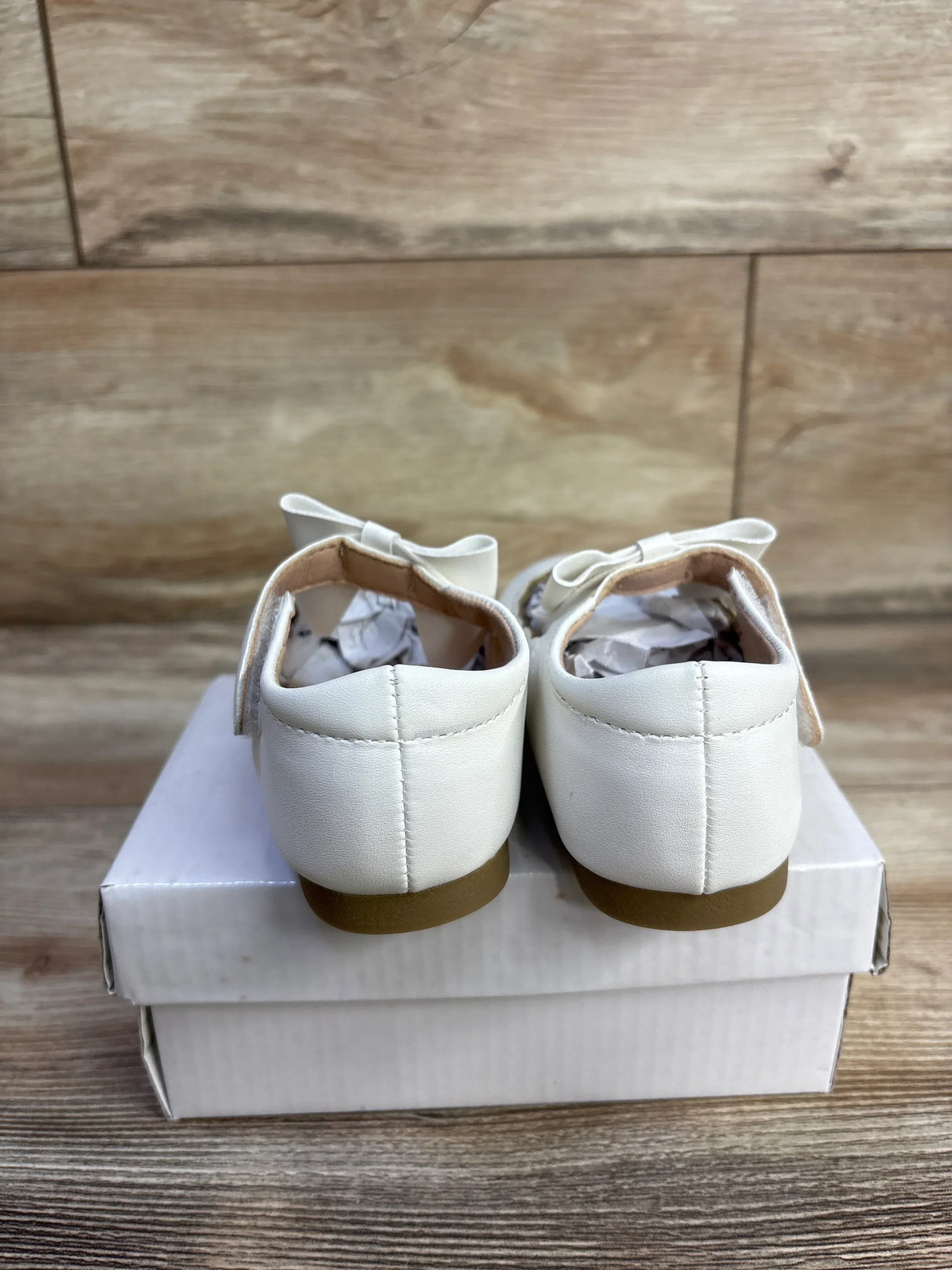 Rear view of white leather baby princess flats size 7C showcasing bows and tan rubber soles on a white shoebox and wood floor