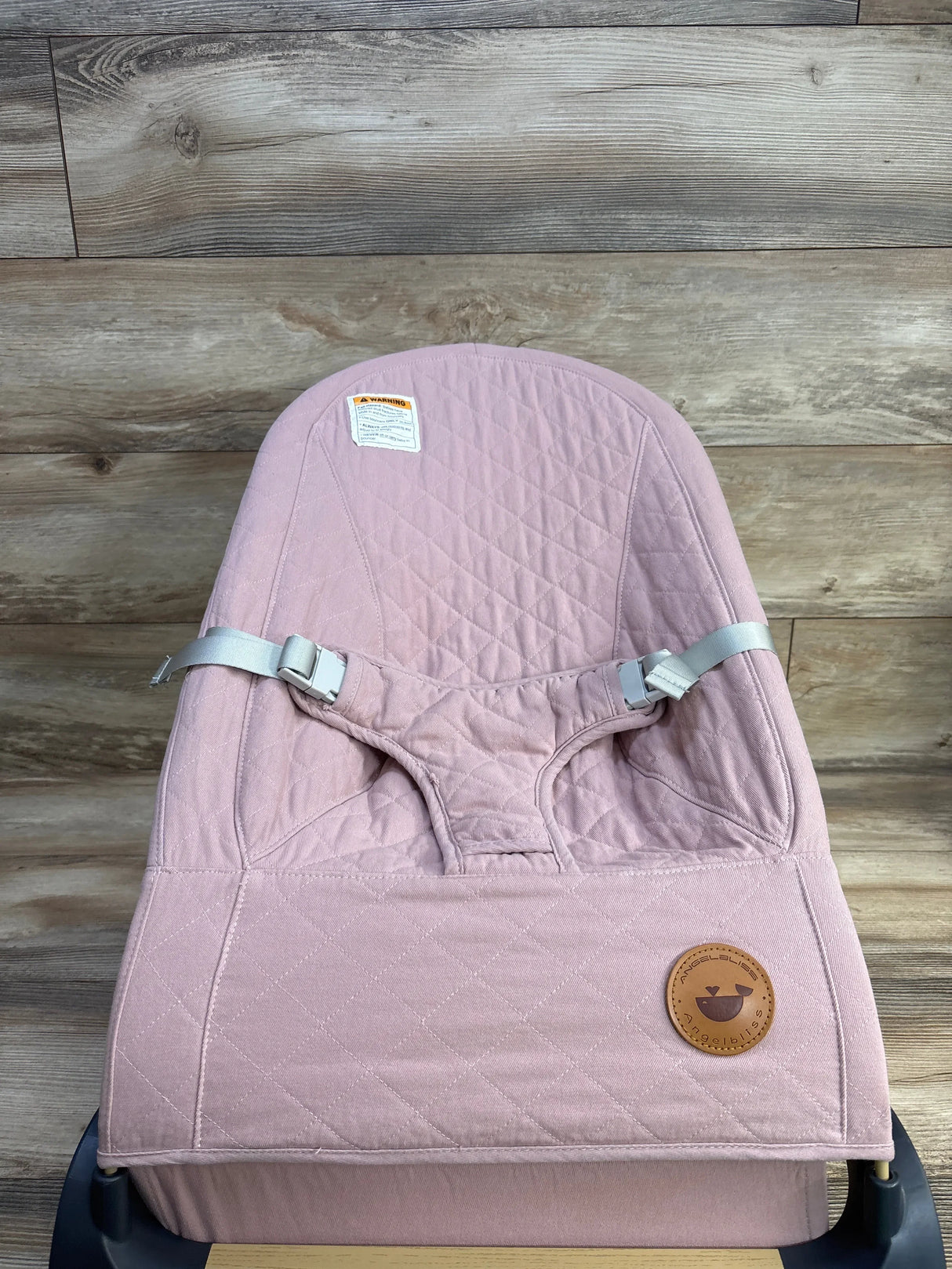 Front view of ANGELBLISS baby bouncer in pink, quilted seat with adjustable gray straps and leather logo on wood background