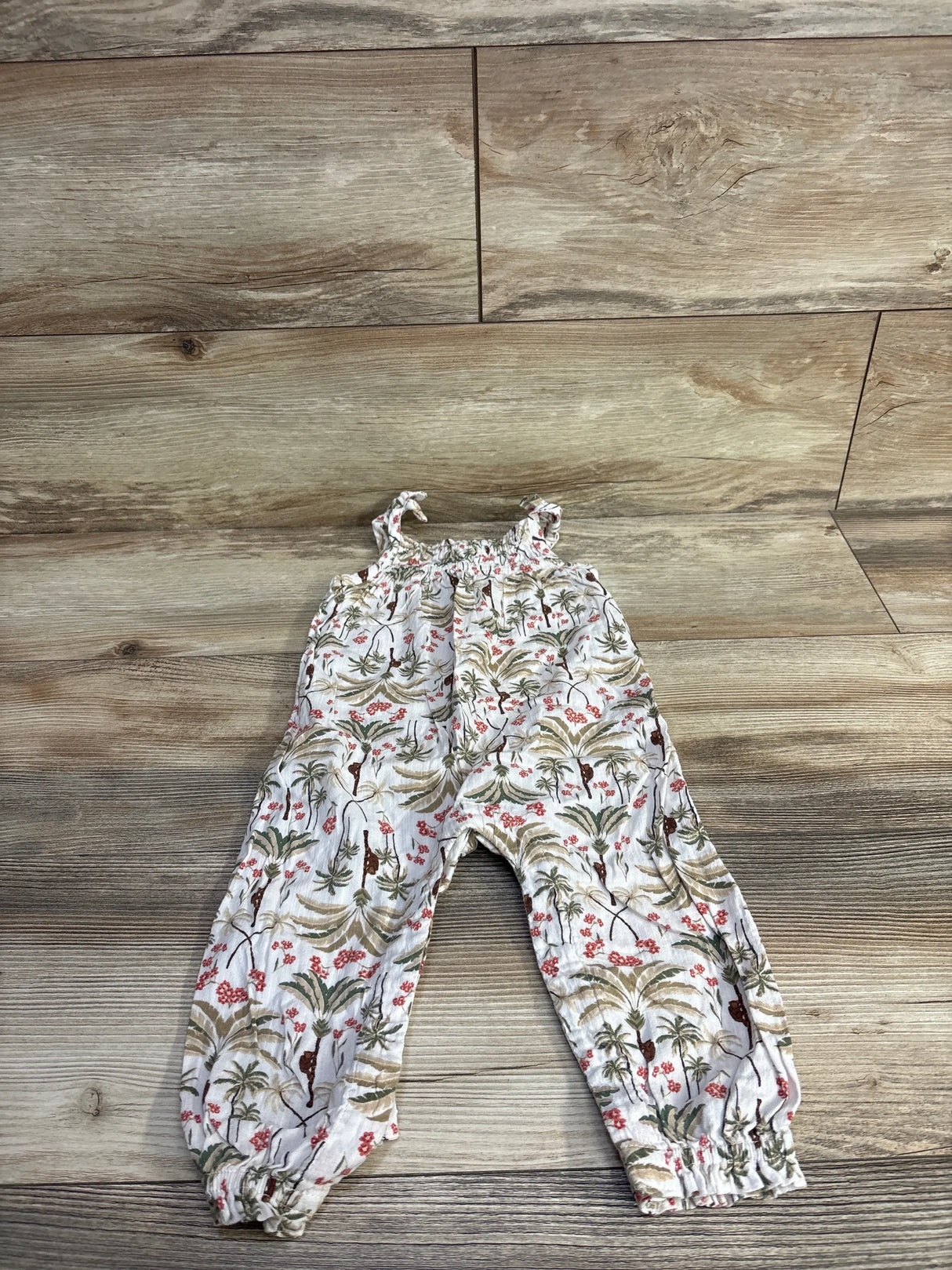Flat lay of cream sleeveless toddler jumpsuit with tie shoulder knots and palm tree floral print on wood background