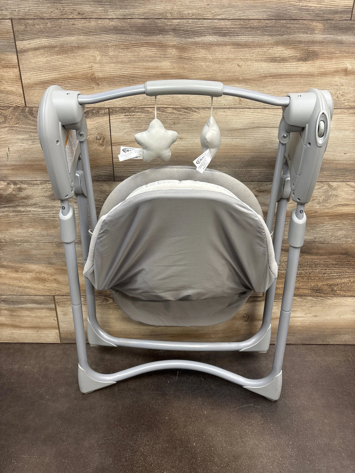 Front view of Graco Slim Spaces Compact Baby Swing in Reign with gray seat, canopy, and hanging toys on wood backdrop