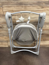 Front view of Graco Slim Spaces Compact Baby Swing in Reign with gray seat, canopy, and hanging toys on wood backdrop