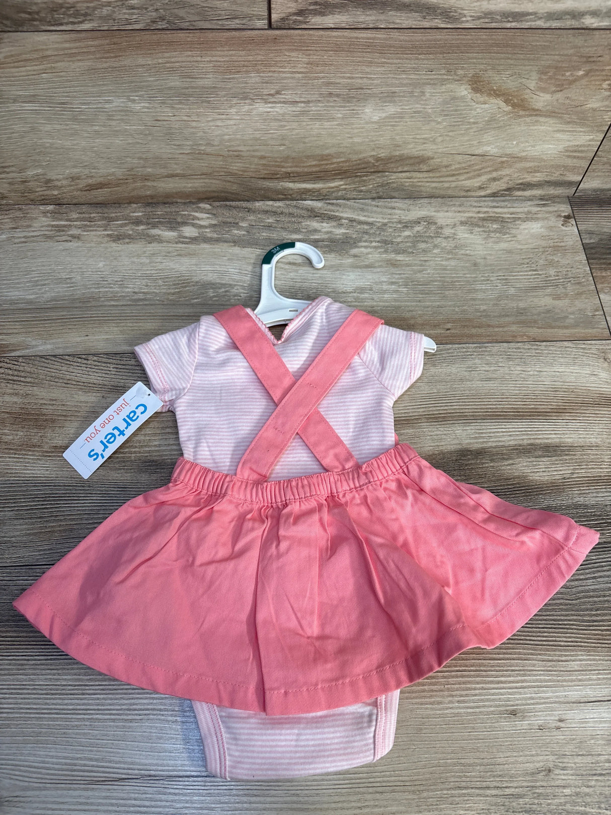Pink baby girl dress with attached skirt and cross straps, displayed on a hanger, wooden background