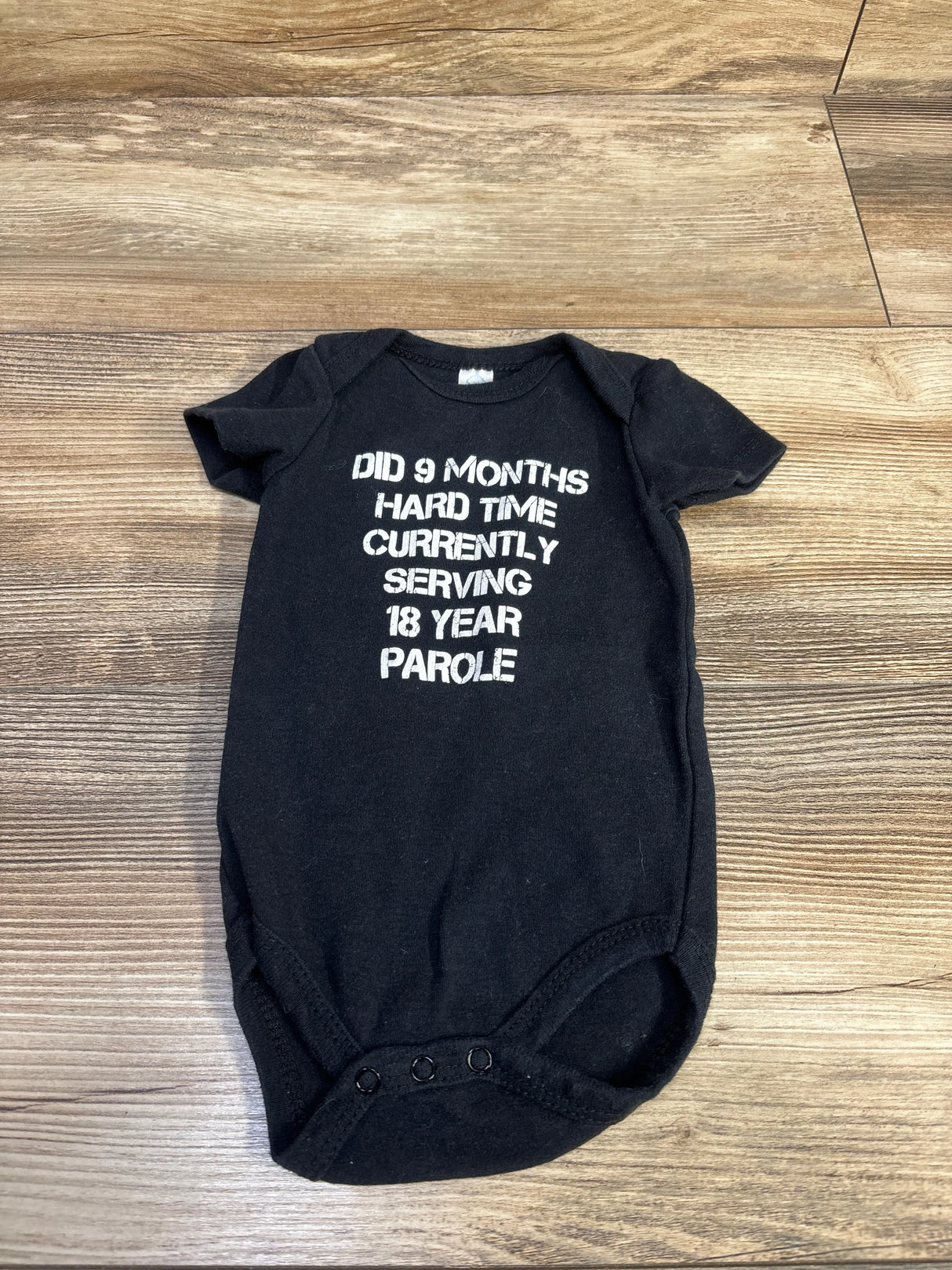Flat lay of black newborn bodysuit showing white text Did 9 months hard time currently serving 18 year parole on wood background