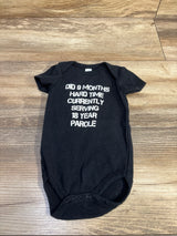 Flat lay of black newborn bodysuit showing white text Did 9 months hard time currently serving 18 year parole on wood background