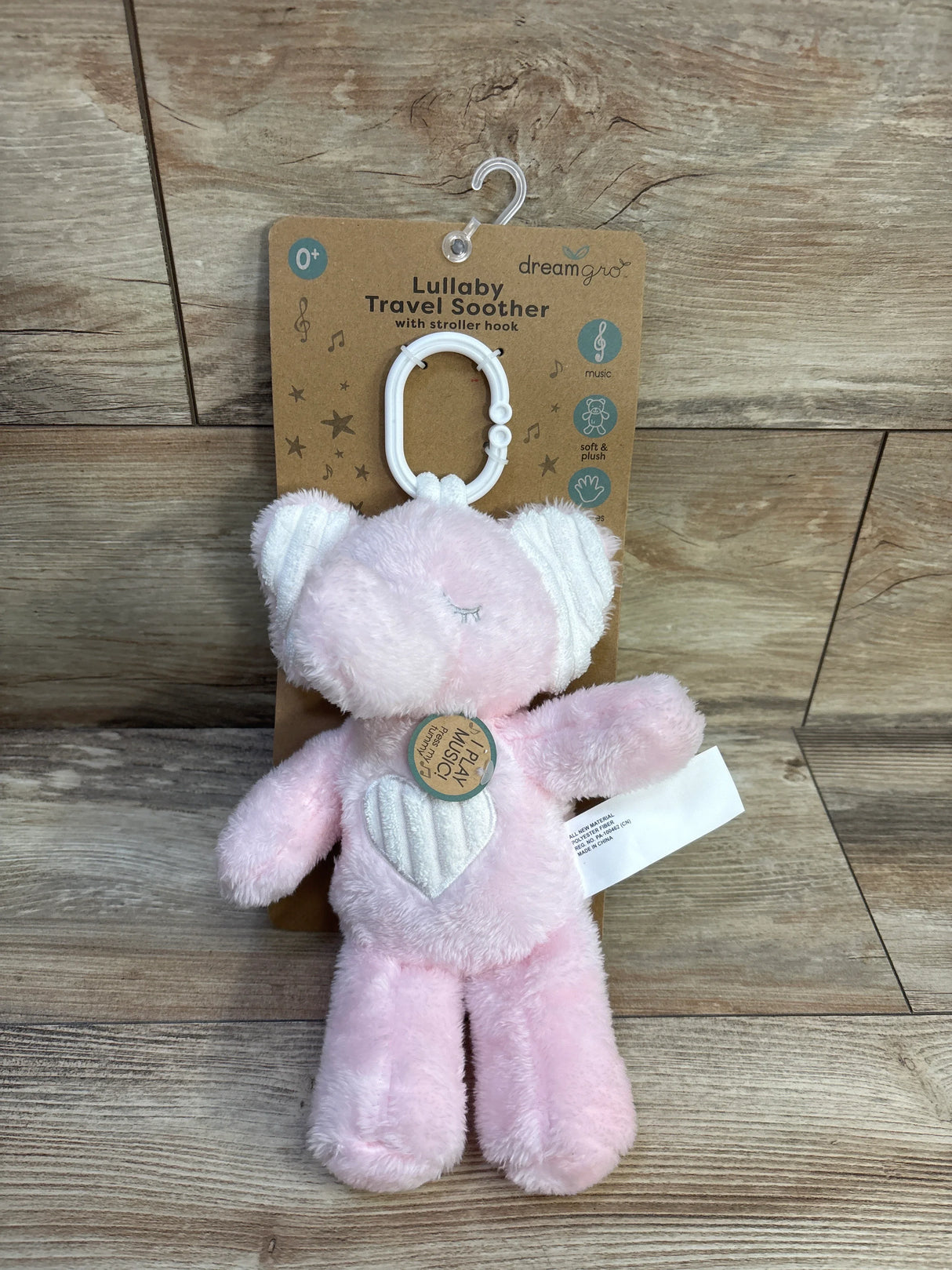 Front view of DreamGro Lullaby travel soother pink elephant showing white stroller hook, heart patch, and soft plush against a wood background
