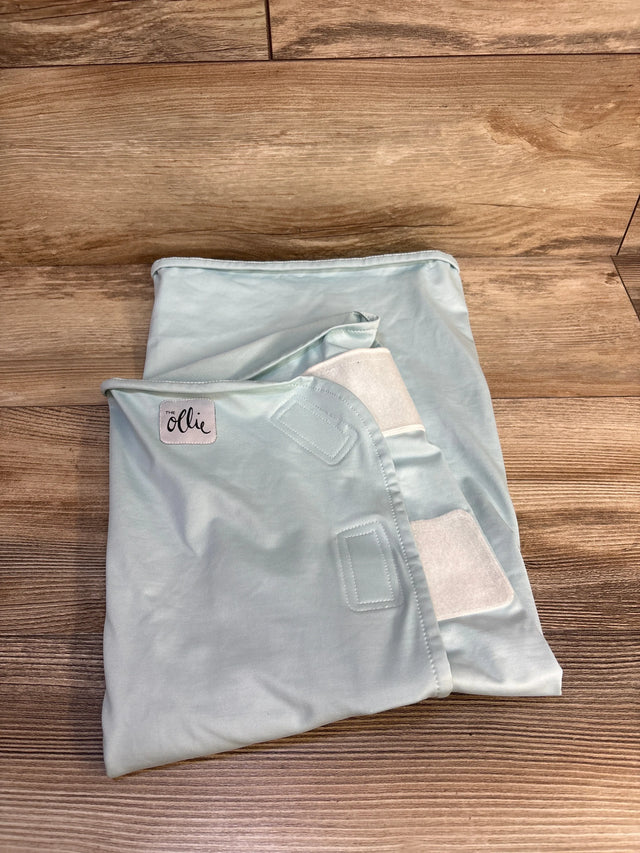 Light blue baby swaddle blanket with 'The Ollie' label, soft fabric and Velcro straps, on wood floor