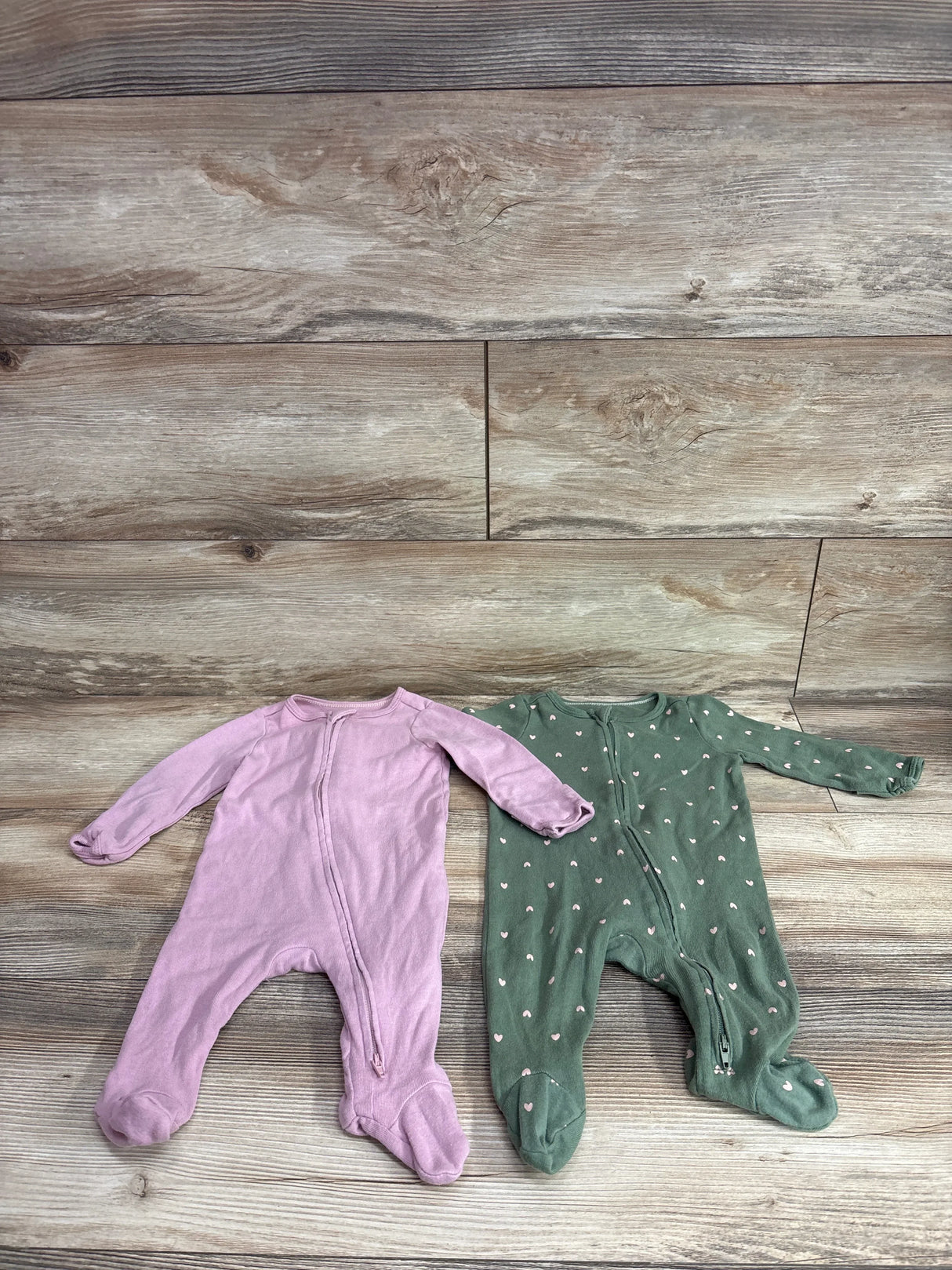 Flat lay of two Cloud Island footed sleepers with front zippers, one mauve and one green with small hearts on wood background