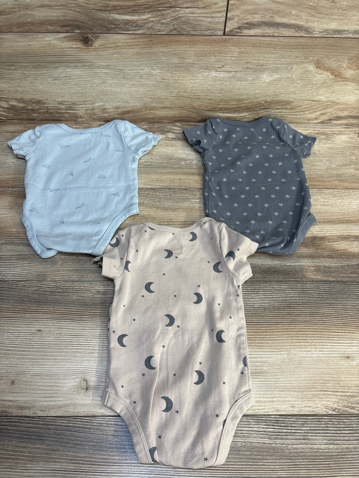 Three baby onesies with moon, star, and paper airplane prints on wood floor, ideal for newborns and infants.