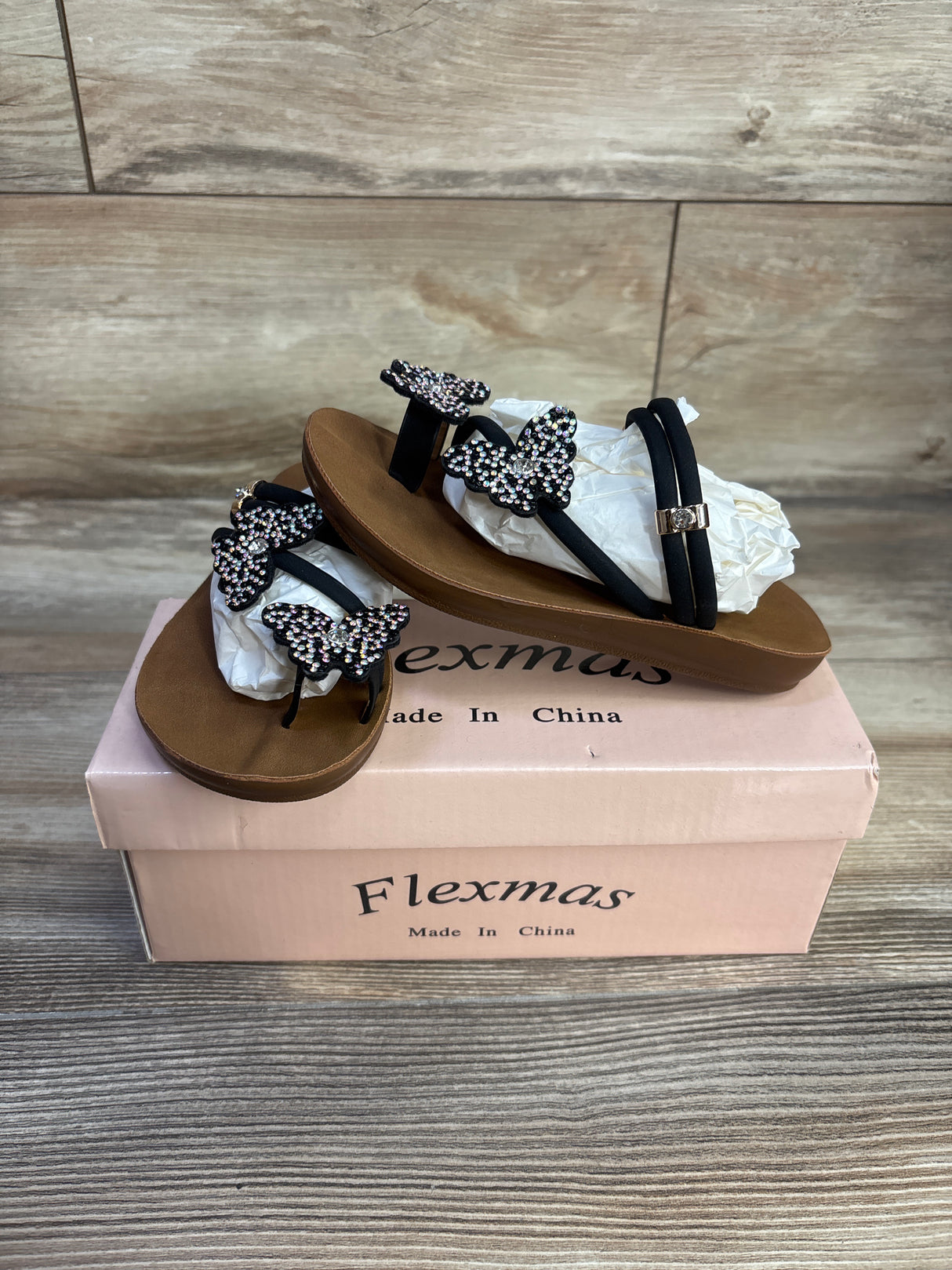 Angled side view of black sandals with rhinestone butterfly straps on a pink Flexmas box on wood surface