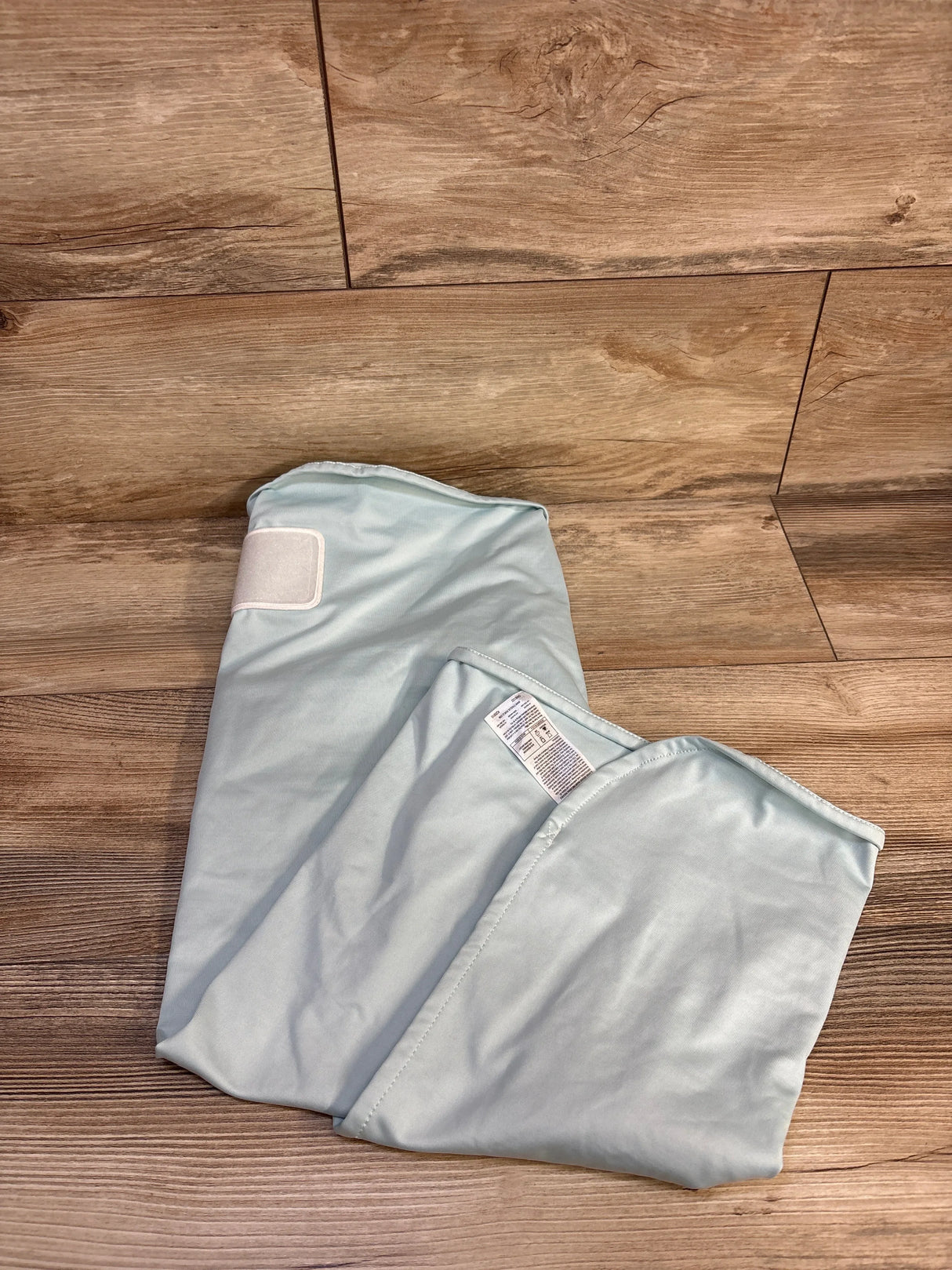 Top-down view of The Ollie World newborn swaddle in light blue showing Velcro closure and care label on a wood floor