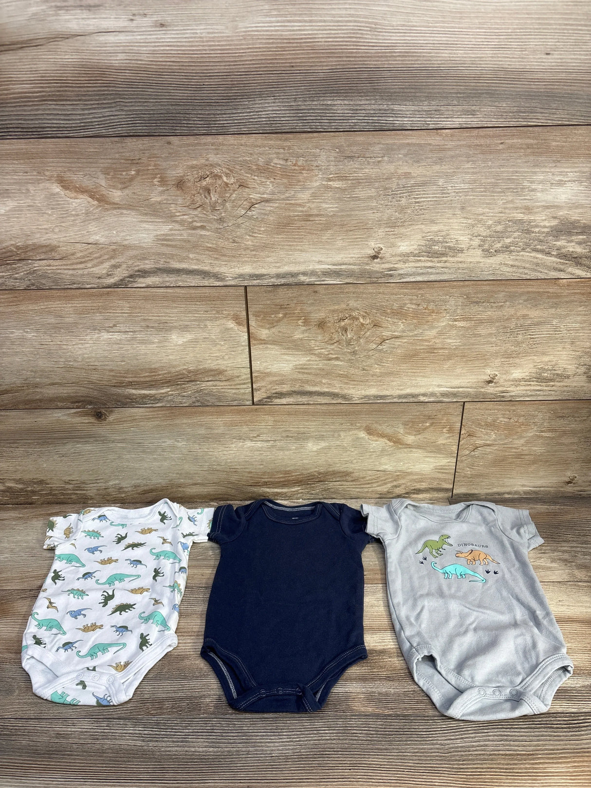 Flat lay of 3-pack Hudson Baby bodysuits size 3-6M showing dinosaur prints and navy bodysuit on wood background