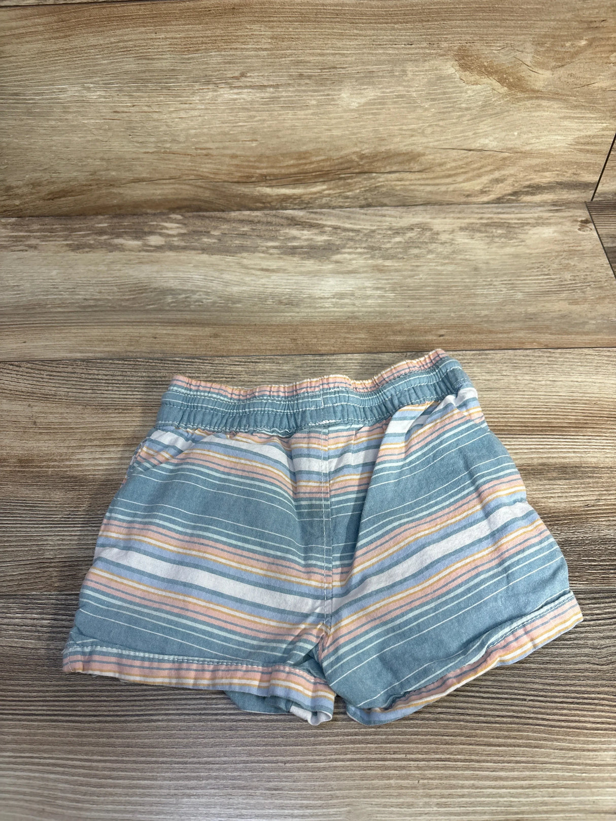 Top view of blue OshKosh cotton shorts with elastic waistband and pastel horizontal stripes on wood background
