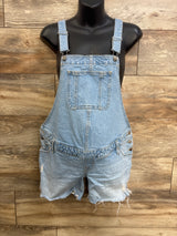Front view of light blue maternity denim shortalls with straps, bib pocket, side buttons and frayed hem on wood