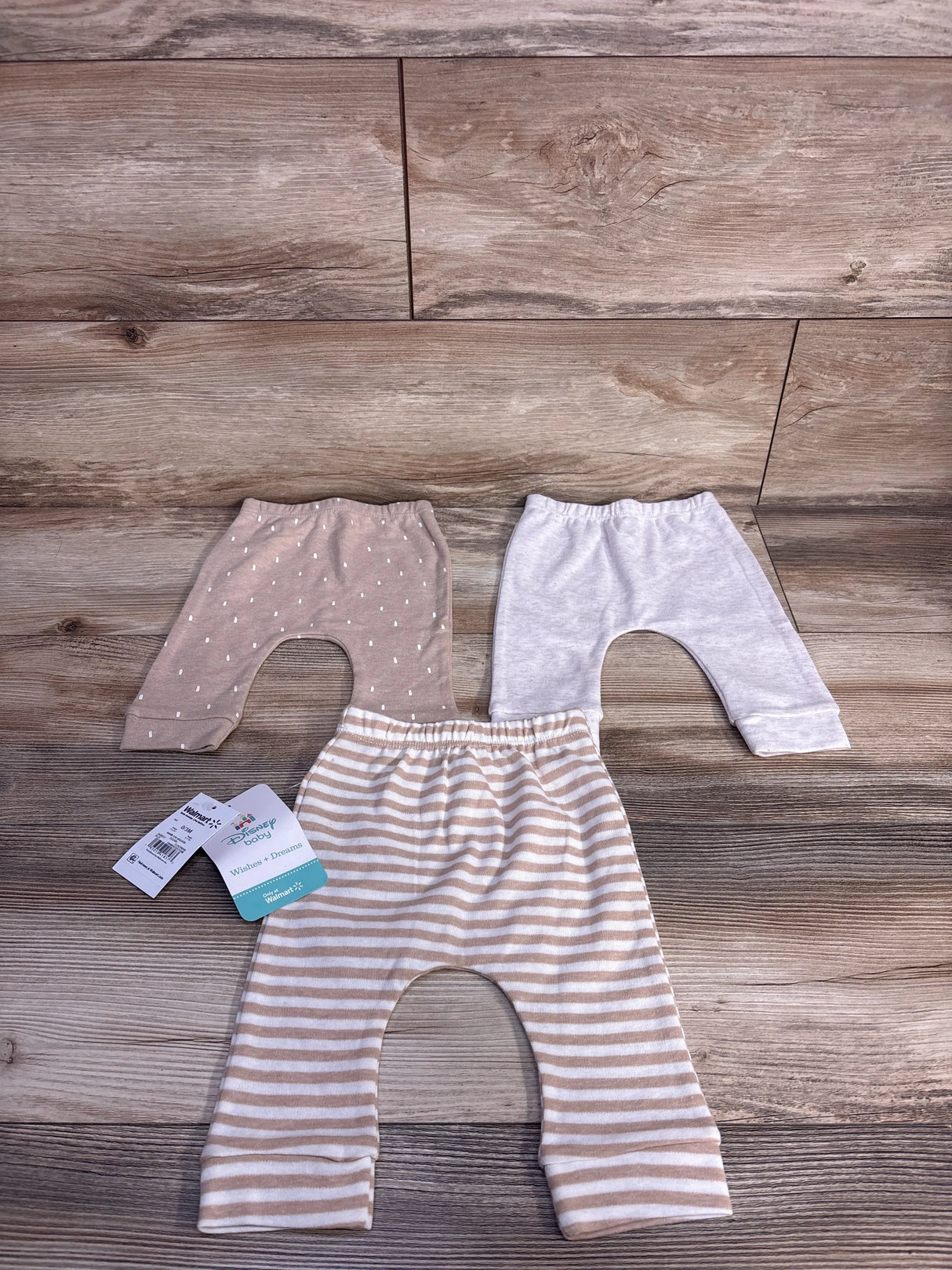 Flat lay of three newborn Disney Baby pants, tan dotted, light gray, and tan-and-white striped, showing elastic waists on wood background