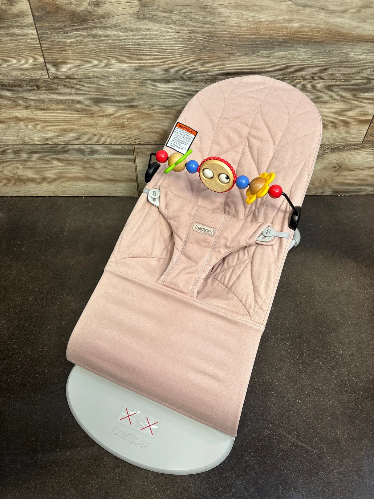 Top-down view of Baby Bjorn Bouncer Bliss in dusty pink, quilted petal seat with wooden-bead toy bar on dark floor
