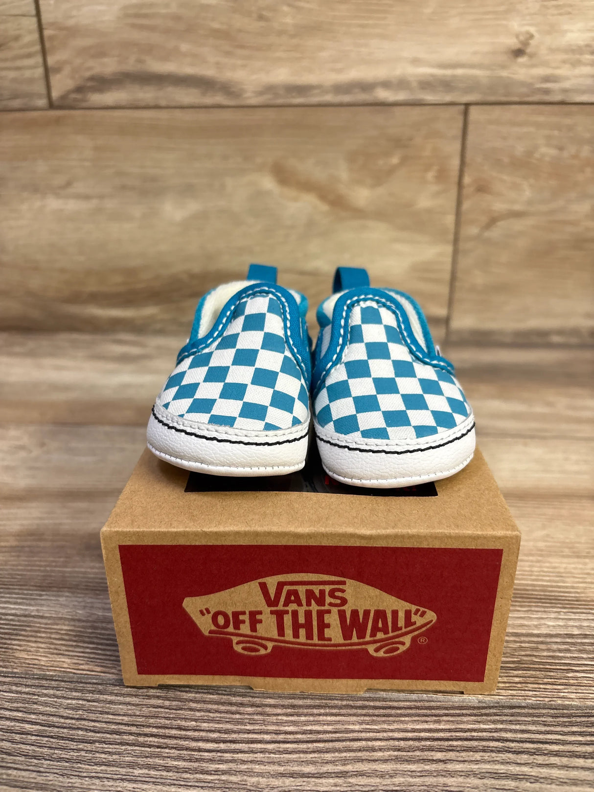 Front view of Vans Slip-On V crib shoes showcasing blue and white checkerboard canvas and white rubber toe on a Vans box