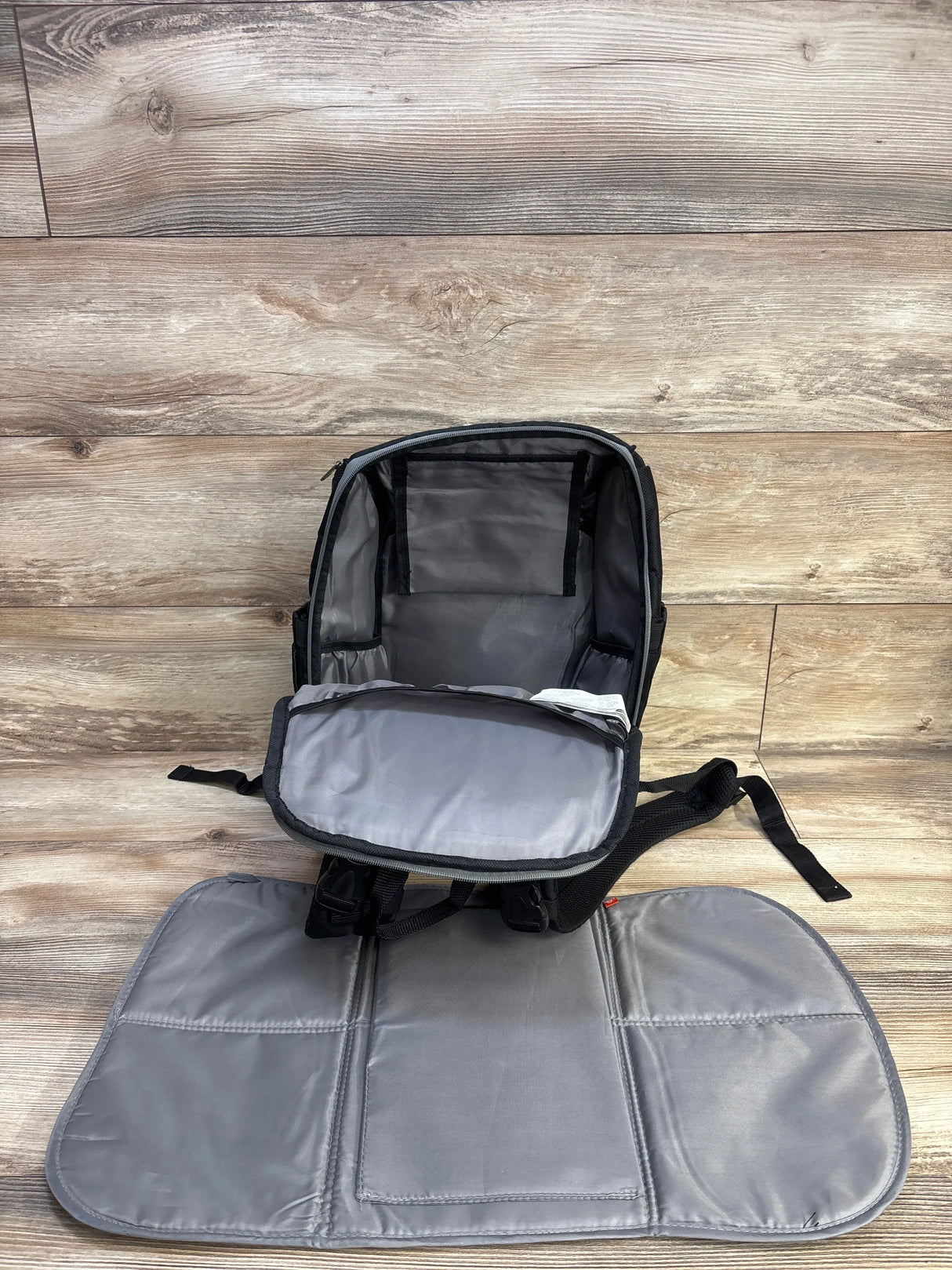 Open view of Kaden FastFinder diaper backpack showing gray interior and fold-out changing pad on wood floor