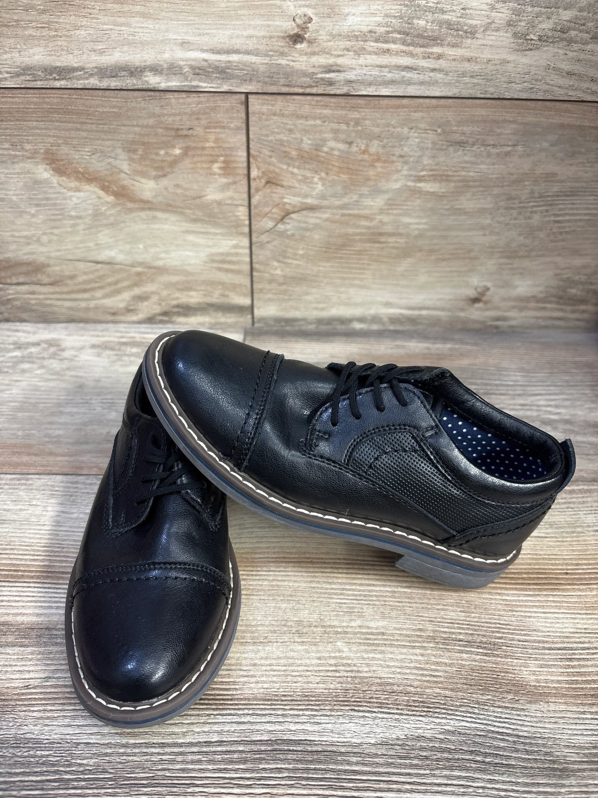 Angled view of black toddler cap-toe oxford by Steve Madden, white welt stitching and polka-dot lining on wood backdrop