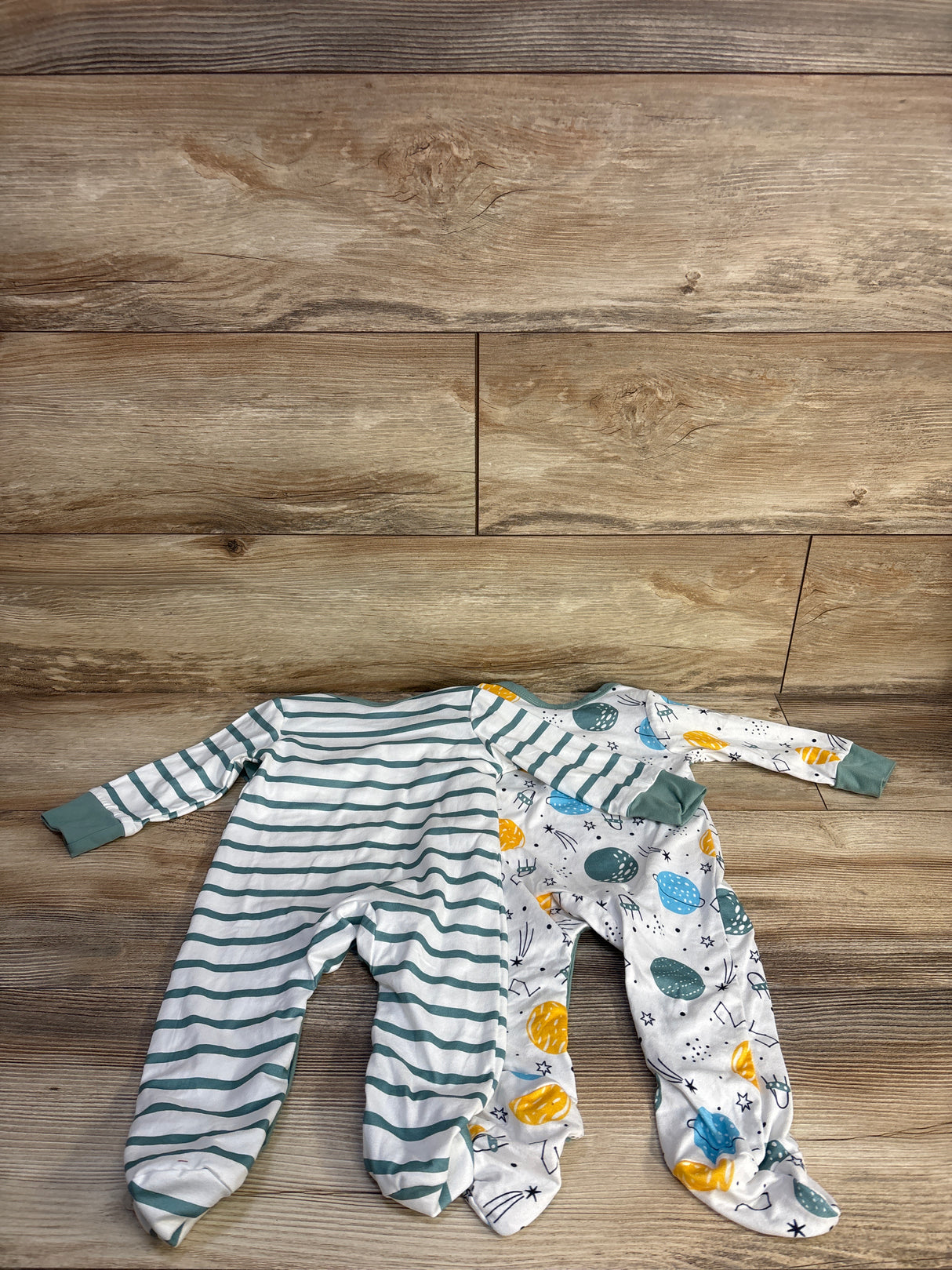 Top view of 2 newborn footed coveralls: teal striped and white with planet print, on wood background