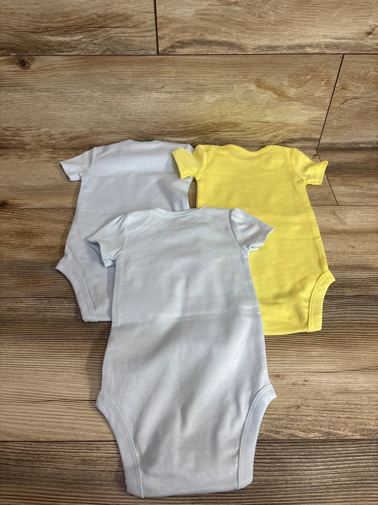 Back view of three infant short-sleeve bodysuits showing two light blue and one yellow, laid flat on wood background