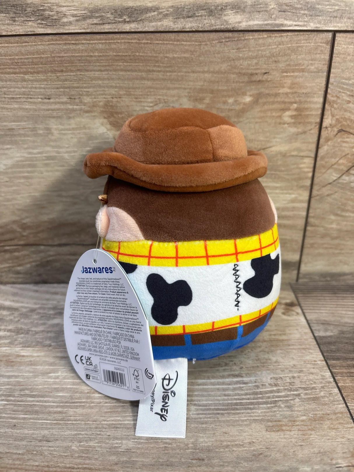 Rear view of 5-inch Woody Squishmallow plush showing brown hat, cow-print vest and Jazwares and Disney tags on wood background