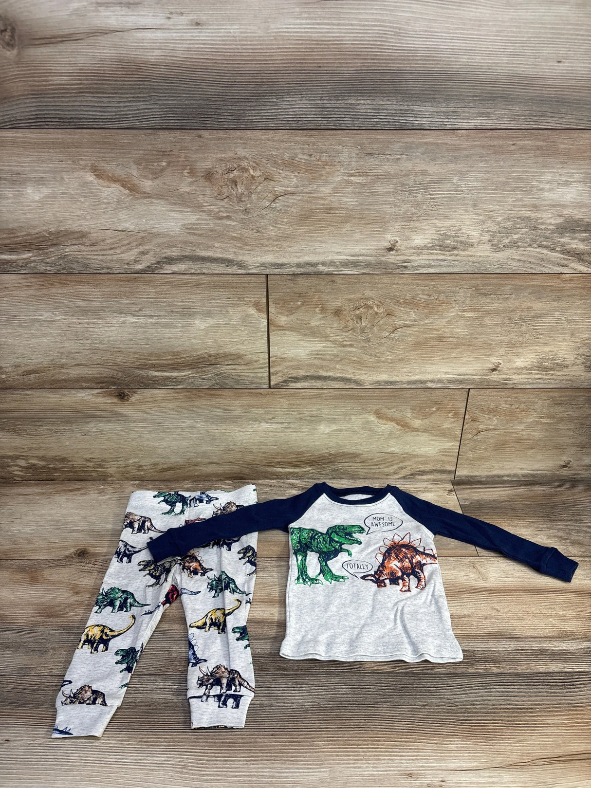 Flat lay of grey 2-piece toddler dinosaur pajamas: navy raglan top with speech-bubble dinosaur graphic and matching dinosaur-print pants on wood floor