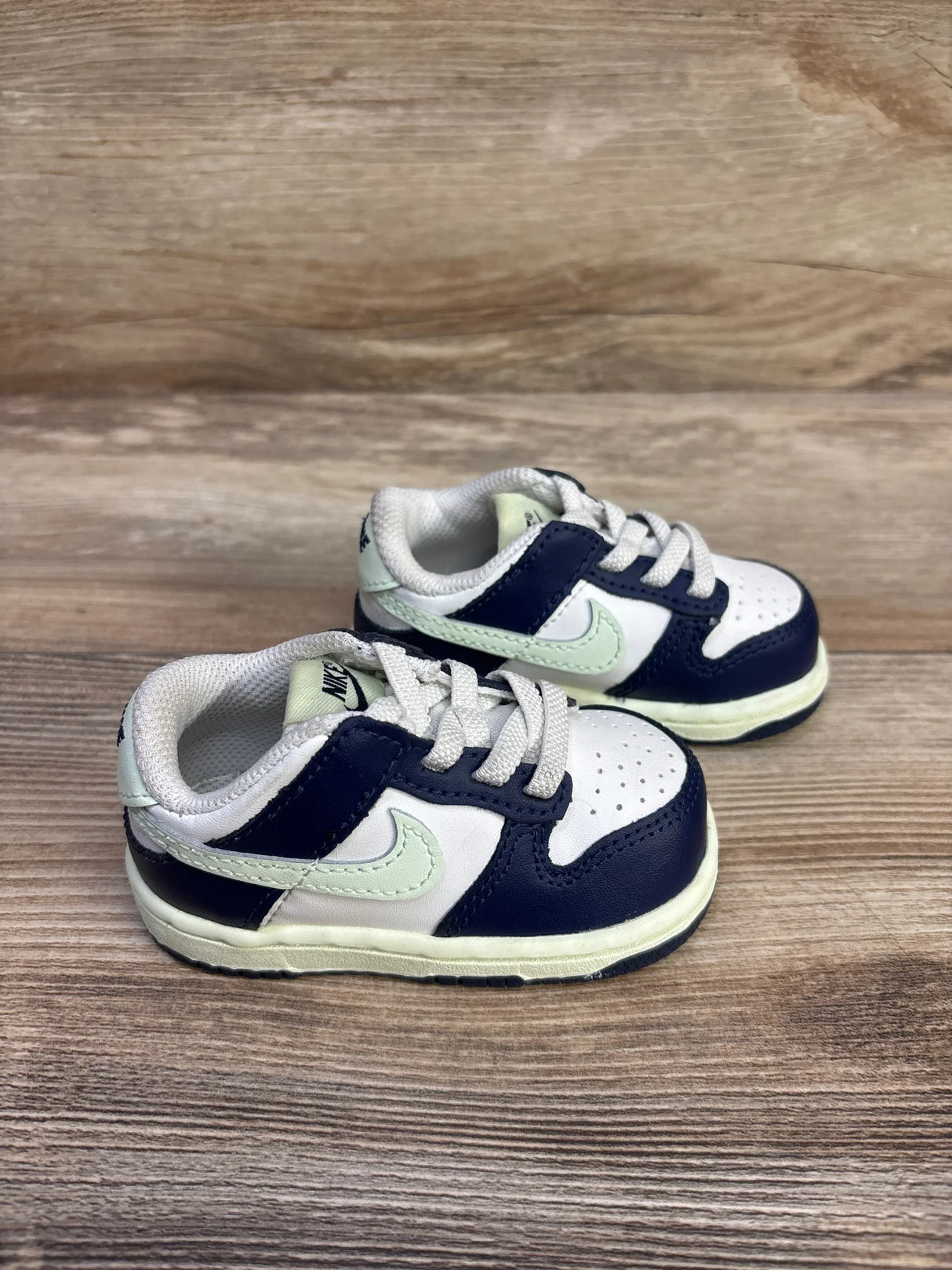 Side view of toddler Nike Dunk Low sneakers in navy and pale green showing perforated toe and laces on wood surface