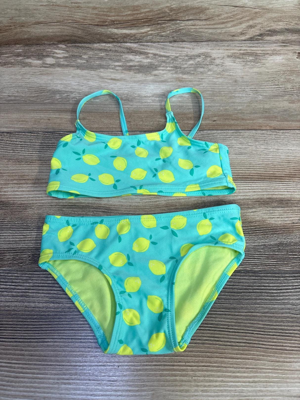 Flat-lay of girls' 2-piece teal lemon-print bikini (2T) showing thin straps and lined full bottoms on wood background