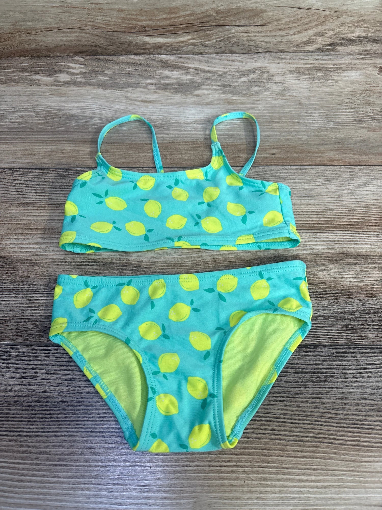 Flat-lay of girls' 2-piece teal lemon-print bikini (2T) showing thin straps and lined full bottoms on wood background