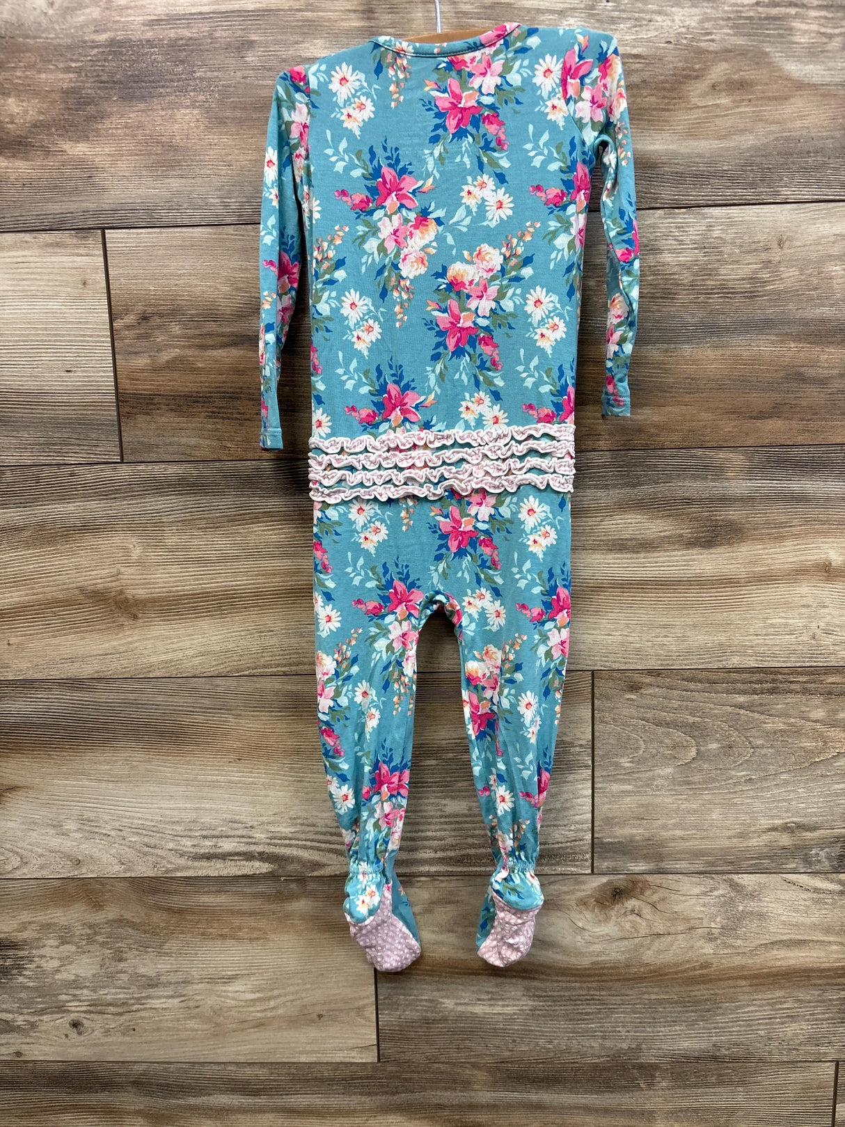 Back view of RuffleButts SoftSnooze footie showing seat ruffles, teal floral print and pink grippy soles on wood backdrop