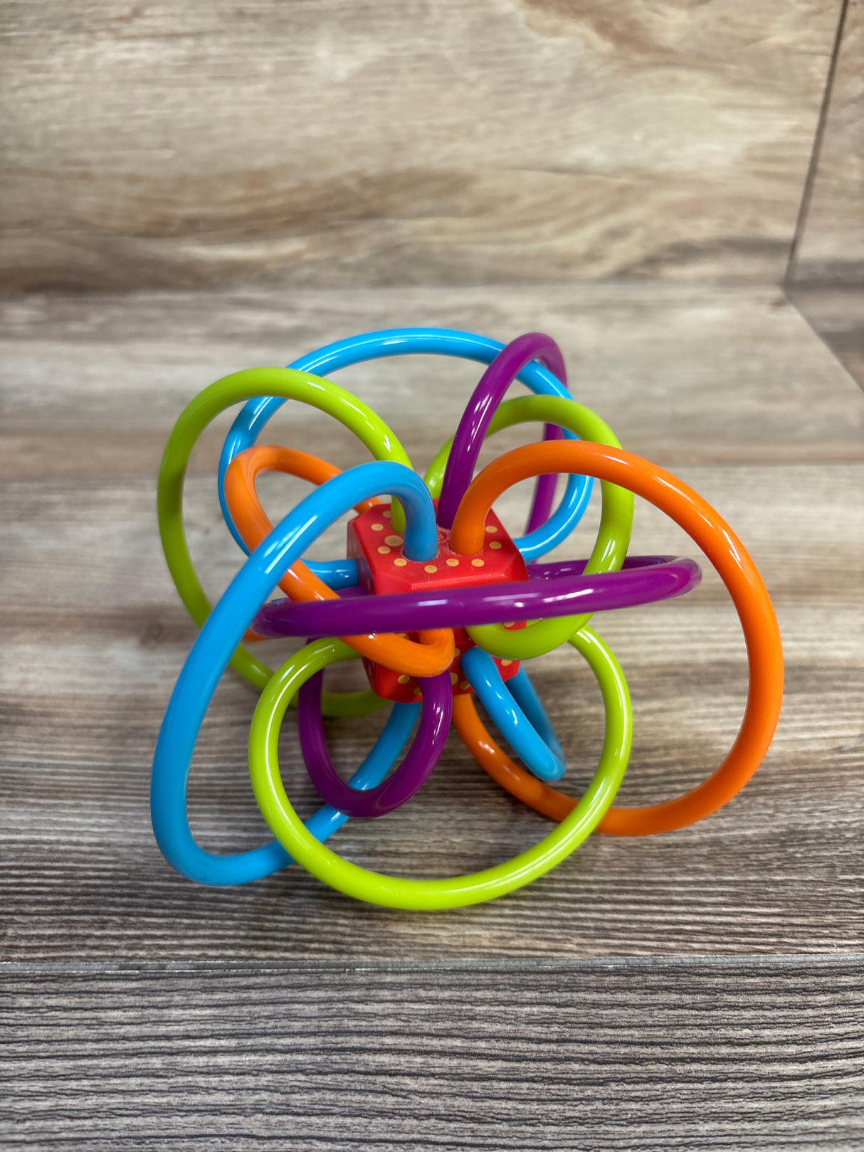 Angled front view of Manhattan Toy Winkel rattle and sensory teether showcasing multicolored flexible loops and red dotted center on a woodgrain surface