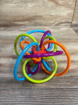 Angled front view of Manhattan Toy Winkel rattle and sensory teether showcasing multicolored flexible loops and red dotted center on a woodgrain surface