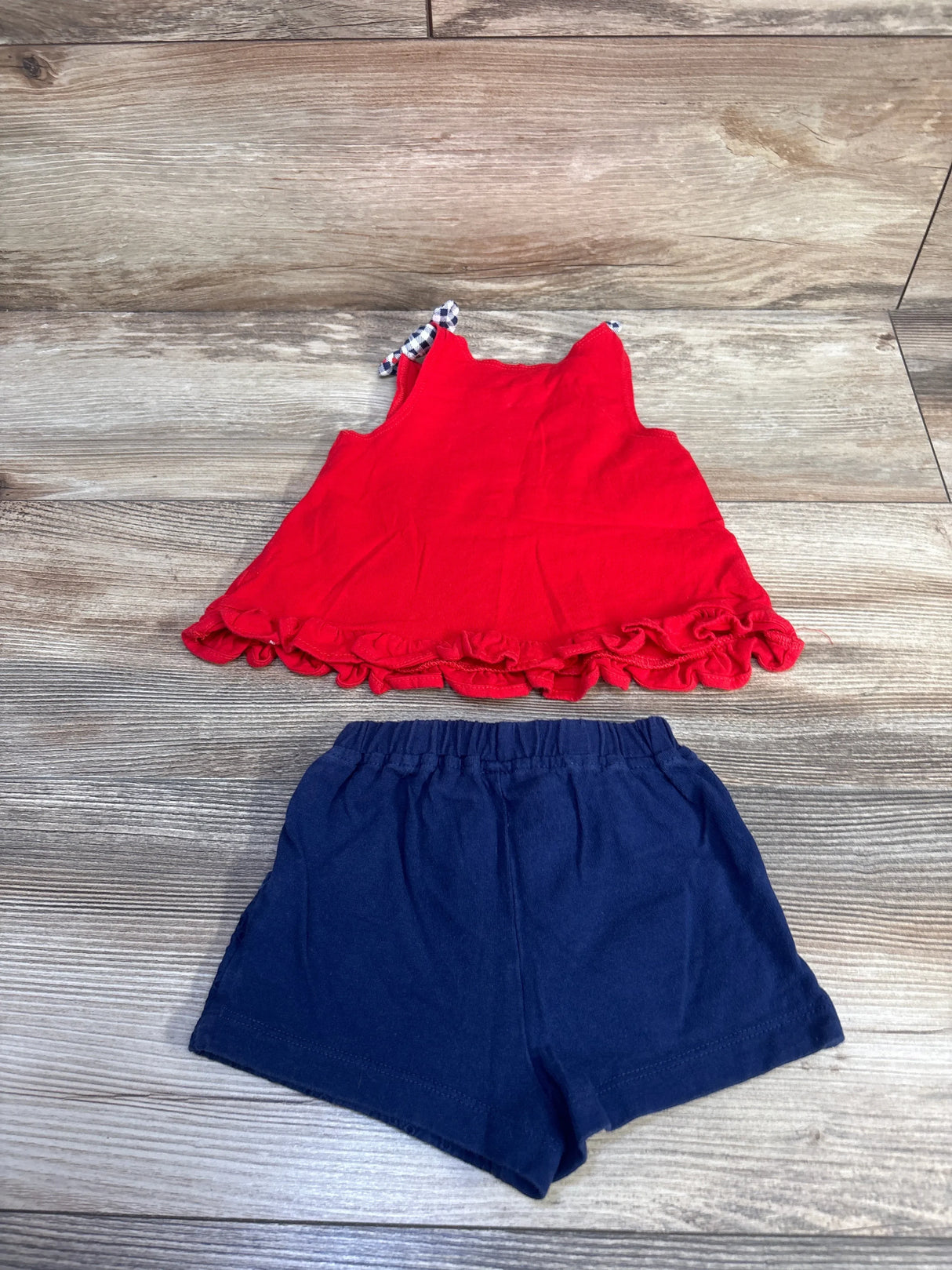 Top-down view of baby red tank top showcasing ruffle hem and gingham tie straps with navy elastic-waist shorts on wood background