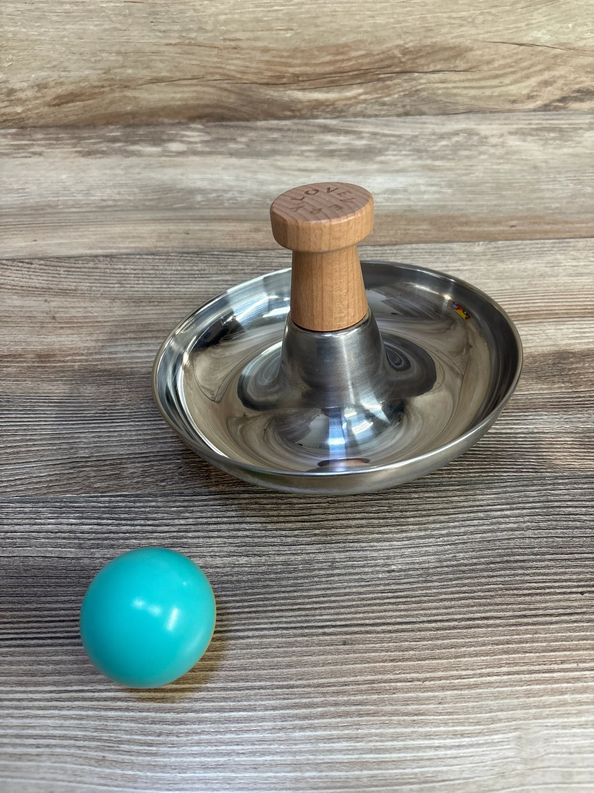 Angled top-down view of Lovevery Stainless Steel Tip & Turn showing stainless steel bowl and wooden knob on a light wood tabletop with teal ball nearby