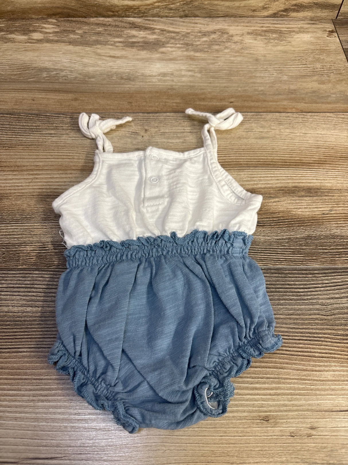 Flat lay of white and blue baby bubble romper, 3M, showing knotted shoulder ties, smocked waist, elastic leg openings on wood