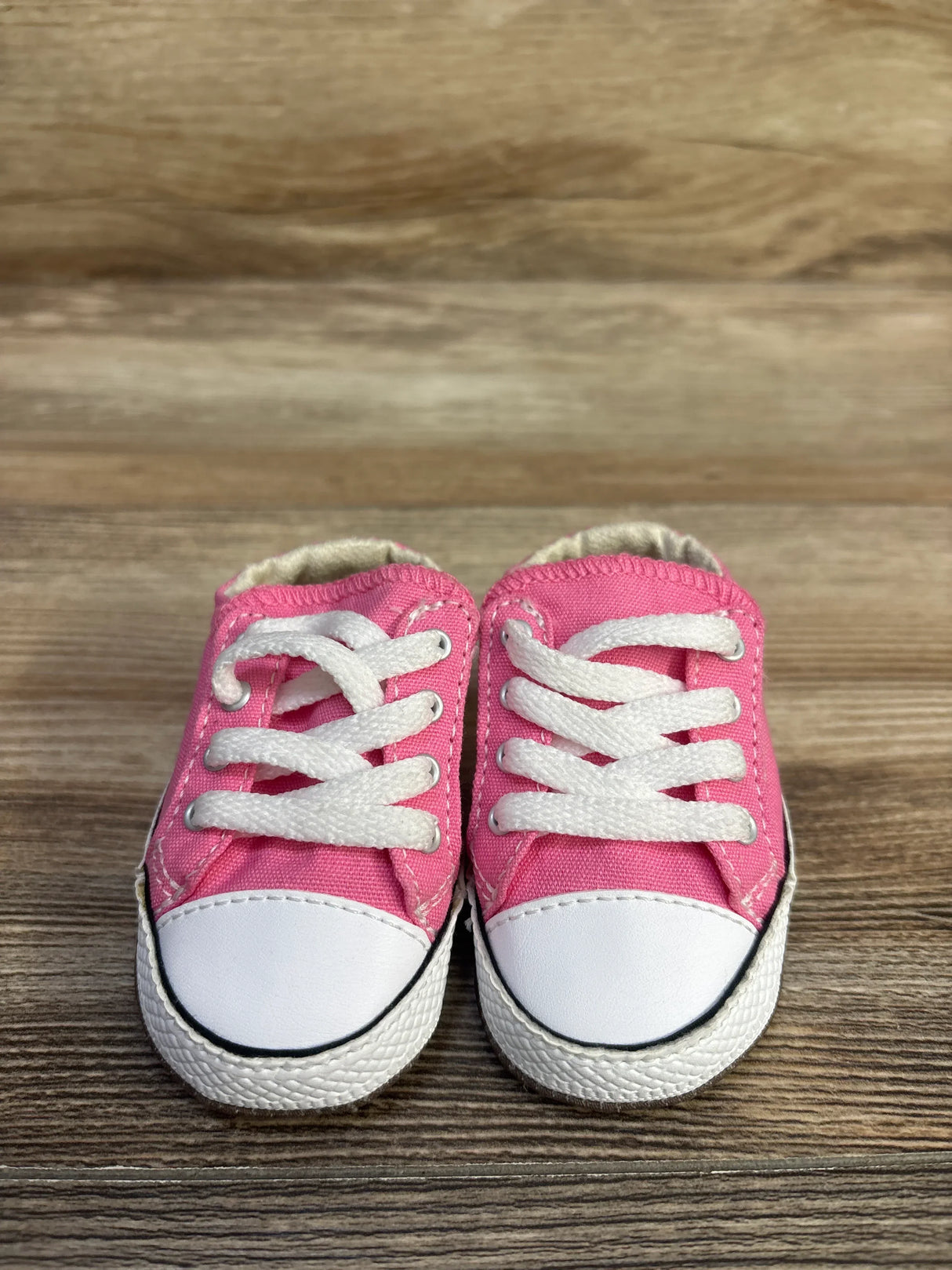 Front view of pink Converse Cribster baby shoes showing white rubber toe caps and laces on wood tabletop